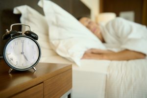 Picture of an alarm clock on a nightstand next to a bed with a woman sleeping in it.