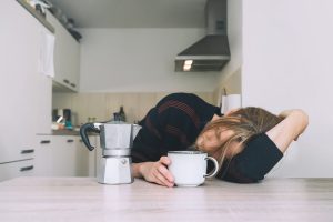 Picture of a woman falling asleep while sitting at a table with a coffee pot and mug in front of her.