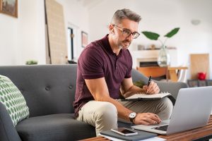 Picture of a man sitting on a couch and writing on a clipboard with a laptop on the coffee table in front of him.